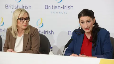 Michelle O'Neill and Emma Little-Pengelly address the press after a British-Irish Council meeting. O'Neill who has blond shoulder length hair and black-rimmed glasses is looking on as Little-Pengelly, who has long black hair, speaks.
O'Neill is wearing a light brown jacket and white shirt, Little-Pengelly a blue jacket and red shirt.