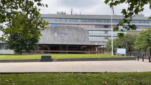 A flat roofed council building made of banded grey concrete and glass with the council's ovaloid chamber protruding to the front, with a grass bank and trees in the foreground 