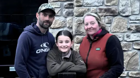 Harry Byatt Three people, a family, stand together in front of a stone wall. The woman, Megan Byatt, is 39 with blonde hair tied back and is wearing a dark red top. The man, Harry Byatt, has a blue top and cap with a black stubble. His daughter, who looks about 11, has a brown top on and dark hair.