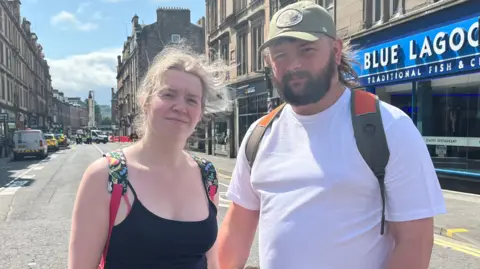 Ellen Fulton, left, staring right at the camera. She has fair hair which is being blown by the wind. She is wearing a black vest top and has the straps of a multi-coloured backpack around her shoulders. Kieran Gibson is on the right. He has a beard and is wearing a khaki-coloured baseball cap. He is wearing a white t-shirt and has the straps of a bag, which are khaki and orange coloured around his shoulders.