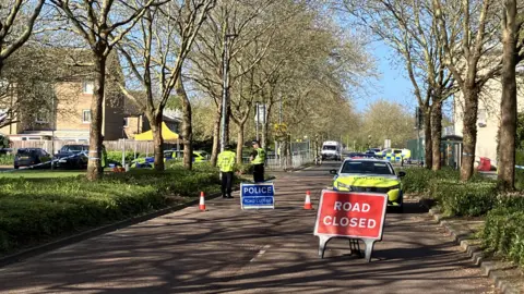 Shariqua Ahmed/BBC A road closure, with two "road closed" signs on the tarmac, a police car and some cones. Two officers are speaking to each other inside the cordon. It is a tree-lined street. A police van and more tape is visible in the background, near the shopping centre entrance.