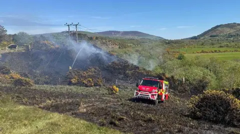 Isle of Man Fire and Rescue Service A charred field with a small hill to the left with electricity lines visible over it. There are gorse bushes near the foot of the hill and a firefighter using a hose. A fire engine is parked in the field and there are rolling hills in the background.