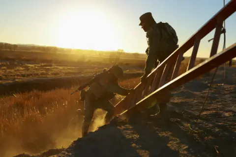 Jose Luis Gonzalez/Reuters Two members of the Mexican National Guard lift a ladder up a slope. One of the guards is holding a rifle. The sun is beating down in the background.