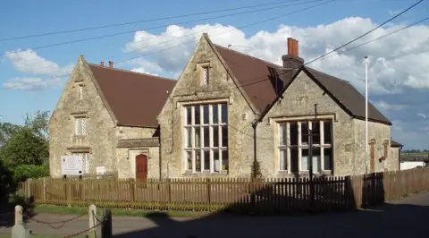 Large stone school buildings with steep gabled roofs surround a fenced outdoor area. There is a telegraph pole to the right. There are clouds in the sky and telegraph cables going to the building. 