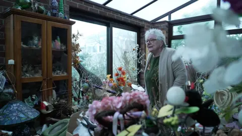 Jayne, who has grey hair and glasses and is wearing a beige cardigan, she is stood in a room full of objects. Behind her, next to her and in front of her are piles of random objects she has collected