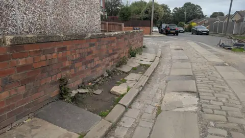 Alex Moss/BBC A residential road which is made up of large, uneven stone slabs and cobblestones, some of which are missing, leaving gaps filled with dirt and gravel. The missing stones create an irregular, broken pattern along the pavement. In the background are several parked cars.