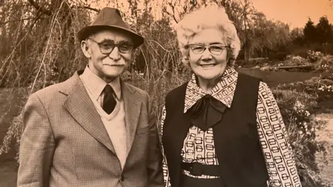 Gooderstone Water Gardens Sepia tinted image of Billy and Florence Knights. He is wearing a hat, smart suit and tie. She is dressed in a white patterned top and black jacket. 