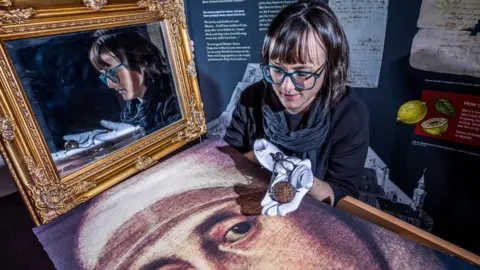 Dr Hannah Thomas holds up with Mary Ward's with portrait of Mary Ward and her Paternoster bead in gloved hands. Dr Thomas wears a grey jumper, dark grey scarf and blue-rimmed glasses. She has short brown hair.