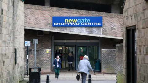 The outside of the shopping centre which is a large brick building with a blue sign above the glass entrance which reads 'newgate shopping centre'. Two people are in the frame, with a man walking towards the camera and a woman walking towards the shopping centre.
