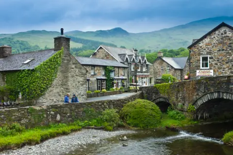 Getty Images A picturesque village is pictured with stone houses and bridge crossing a river. Ivy covers some of the buildings, with mountains in the background. 