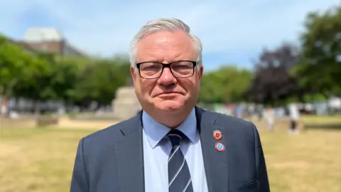 Constable Richard Vibert is standing in Parade Park. He is looking into the camera. He is wearing a navy suit with a blue shirt and striped navy tie. He has parish badges on his right lapel.