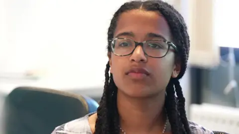 Alyssia, 17, is wearing glasses and has long braids. She is sitting at a table taking part in a discussion 