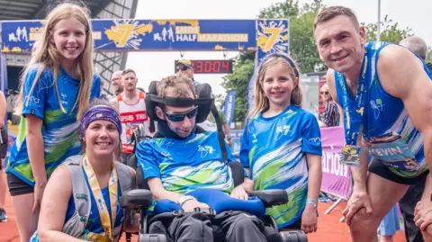 PA Media A group picture of a smiling woman knelt down next to a man in a wheelchair. They are surrounded by two smiling girls and a smiling man who is standing up. The woman and standing man are both wearing medals. In the background there is a blue and yellow coloured arch indicating the end of the Leeds Marathon.
