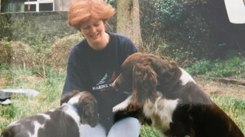 SARAH BLACK Shona Campbell in her 30s with short ginger hair as she kneels on the grass playing with two brown and white springer spaniels.