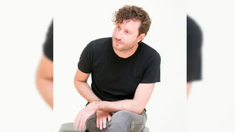 Will Hammond, director of the School of Performance at LIPA, sits on a chair looking out to the left. He is wearing grey jeans and a black t-shirt.