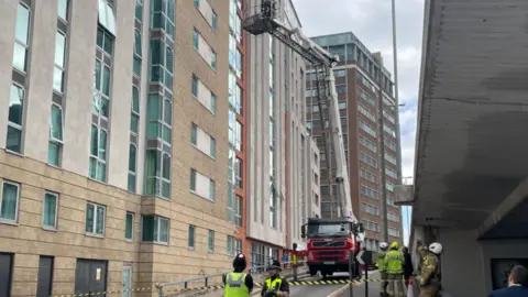 BBC Police and fire officers stood on a street that is taped off, as a fire engine with a hydraulic platform is parked next to high-rise buildings. Two fire officers are on the platform inspecting a building. More buildings can be seen in the background.