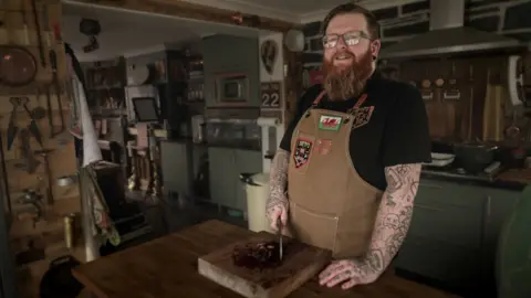 Matt O’Brien standing in his kitchen holding a chef's knife over some meat he has been chopping