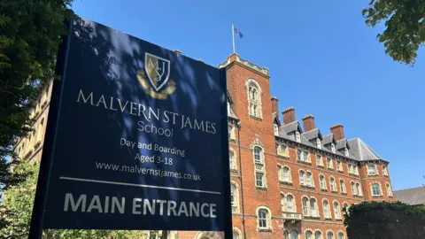 A five-storey red brick school building with a dark blue sign in the foreground with white writing that identifies the school as Malvern St James School. It is a day and boarding school. The image is set against a blue sky and there are trees overhanging part of the photo.