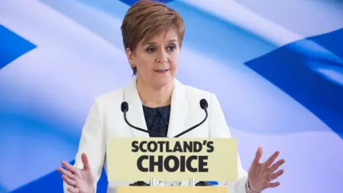 PA Media A woman with short fair hair speaks at a podium in front of a saltire. Wearing a white suit and dark top, she is holding her hands out in front of her. 