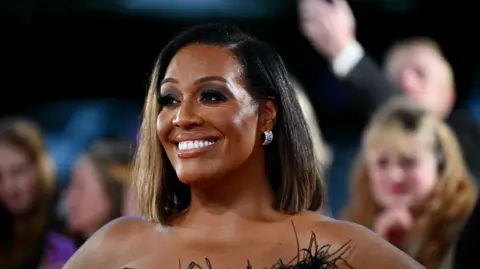 A close-up image of Alison Hammond on a red carpet. She grins into the distance, wearing silver earrings. The top of her dress, which has black feathers, is just visible. The background is blurred but several people in the crowd behind her are visible.