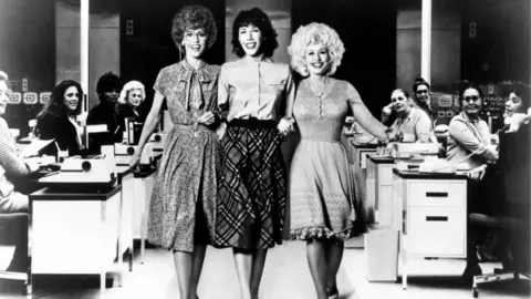 Getty Images A black and white image shows a film still from 1980 comedy film 9 to 5, with Dolly Parton, her blond hair framing her face over a form-fitting dress, locking arms with actress Lily Tomlin, wearing a light-coloured blouse and dark-patterned skirt, and actress Jane Fonda, wearing a button-up dress with a scarf and glasses, as women sit at their desks looking on in an office