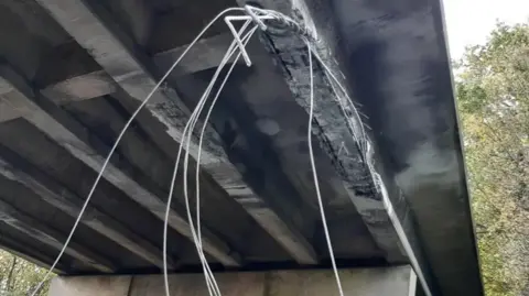 National Highways Damage to the underside of a a bridge caused by a vehicle scraping it. Long metal cables protrude.
