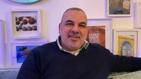 BBC Smiling, bearded man sitting in front of art and wearing a blue jumper and striped shirt