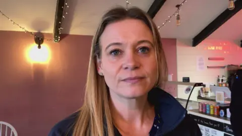 BBC/Jack Fiehn A blonde woman wearing a dark blue jacket is standing in front of a café counter.