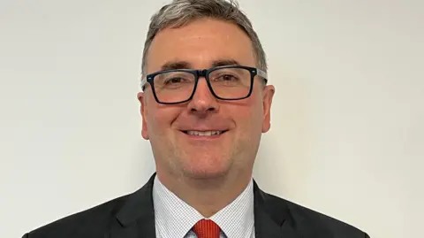 Tim Johnston, a man with grey hair and large black-rimmed glasses, with a bright smile. He wears a dark suit with a light short and red tie.