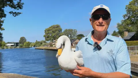 A man with a blue shirt, baseball cap and sunglasses. He is holding a toy swan.