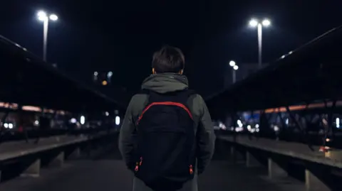 Northamptonshire Police Fire & Crime commissioner A boy with his back to the camera wearing a backpack. It is night time and he appears to be at a train station.