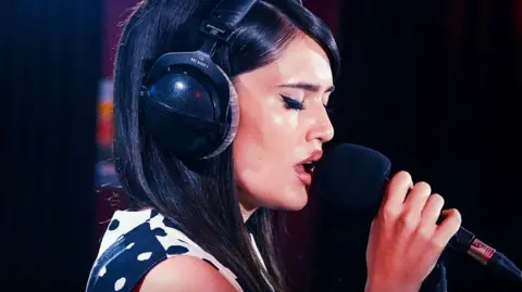 Sienna Spiro sings into a mic, wearing headphones. She has long dark hair and wears a black an white polka dot dress