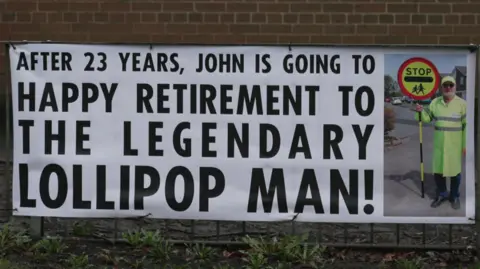 A sign put up on a red-brick wall over some railings that shows a picture of the lollipop man holding his sign, with the message "After 23 years, John is going to stop. Happy retirement to the legendary lollipop man". 