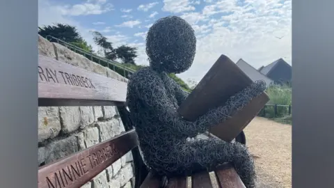 Lyme Regis Town Council Reader sitting on a park bench reading a wooden book