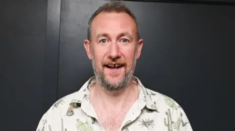 Alex Horne is smiling against a black background. He is wearing a shirt featuring lots of different types of cactus.