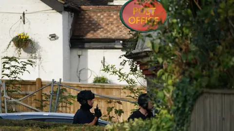 Police officers dressed in black at a Post Office. One holds a radio and the other is partially obscured by greenery. 