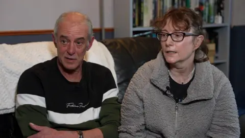 Ken and Kerstin Rudd. Ken is wearing a blue and white striped jumper and has short grey hair. Kerstin is wearing a fluffy grey top over a black t shirt and has glasses and short brown hair. Both are seated on a sofa.