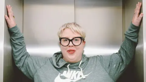 Marea Stamper, aka The Blessed Madonna, poses successful  a assistance   astatine  BBC New Broadcasting House, 2022. She is wearing a greenish  hoodie, has abbreviated  blonde hairsbreadth  and achromatic  glasses.