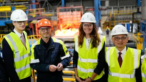 A group of three men and a woman stand in what appears to be a manufacturing plant. They all wear hi-vis jackets and helmets.
