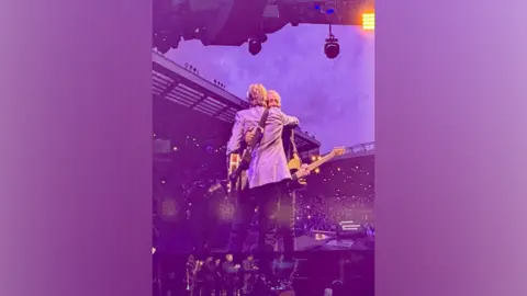 Heart of Liverpool Tours Sir Paul McCartney and Bruce Springsteen hug each other as they hold guitars on stage.