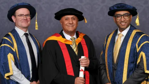 University of Wolverhampton Ranjit Singh Dale in the centre is wearing graduation regalia - a black gown and cap - and is stood in between university representatives