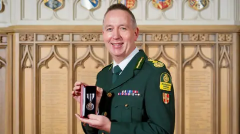 Mark Mapp is standing in uniform smiling at the camera as he holds his Kings Ambulance Medal. It is silver in colour and has a striped ribbon and sits in a presentation box. Behind Mr Mapp is a historic-looking wall covered with etched names, stone reliefs shaped with Gothic arches and brightly painted shields.