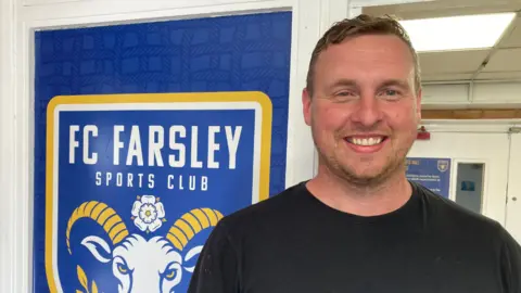David Stockdale is wearing a black t-shirt and is stood in a doorway beside a coat of arms style sign with Farsley FC written on it
