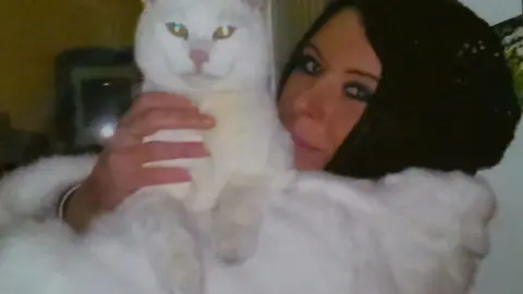 Mandy Hobbis A woman with dark hair wearing a fluffy white jumper, holds a white cat on her chest.