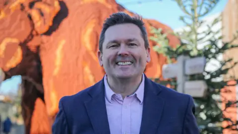 Phil Maclean, a man wearing a pale purple shirt and a dark-coloured suit. He is standing outdoors in front of a large Lego construction of an elephant.