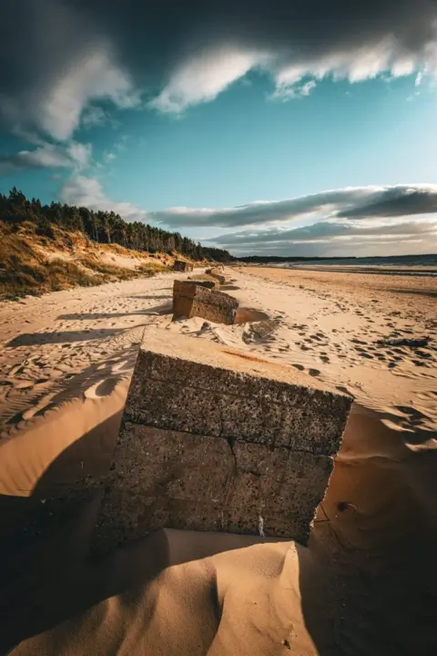 Graham Mackenzie A beach pictured during the day with a cloudy blue sky. There are large stone blocks on the sand stretching into the distance