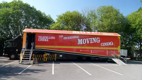 A lorry parked in a car park in front of some trees. The red lorry has the words 'travelling cinema' and 'the incredible moving cinema' on the side. A set of stairs up to a door into the lorry.