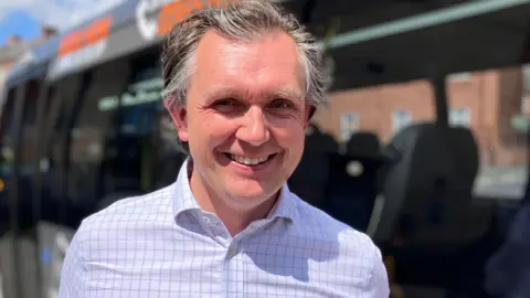 A grey-haired man wearing a white shirt with blue squares, smiling at the camera in front of with windows of a minibus. 