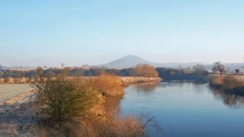 Getty Images A wintery photograph of a river, with the Wrekin hill rising up in the distance, slightly obscured by cloud. The banks of the river are lined with trees and shrubbery, and the grass has a layer of frost on it. The sky is blue and slightly pink, suggesting the photo is taken near sunrise. 