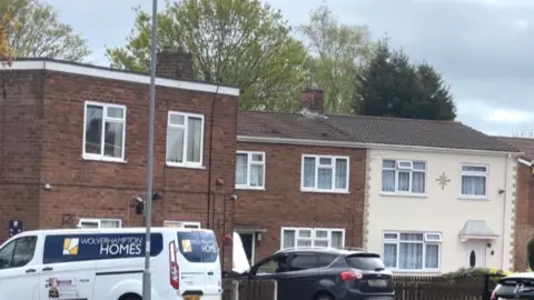 Vehicles are parked on the drives of two homes. One is a van for Wolverhampton Homes. The two-storey square buildings are red brick with another house painted cream.
